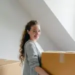 Side view of cheerful female in cardigan with wavy hair holding big cardboard box and looking at camera in attic style living room